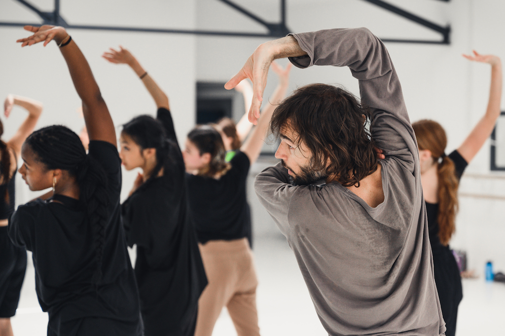 Dancers practice in a studio, lifting their arms above their heads in a posed position